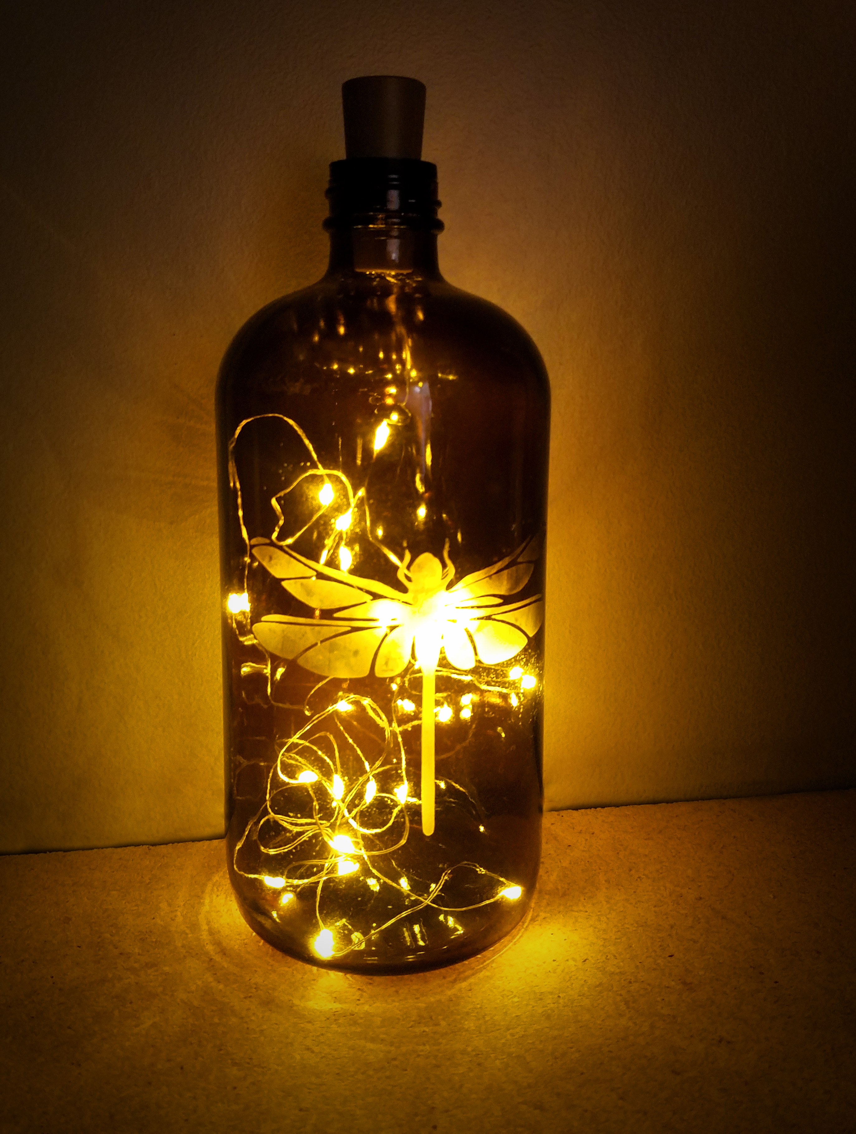 Brown glass bottle with etched dragonfly and string lights glowing inside