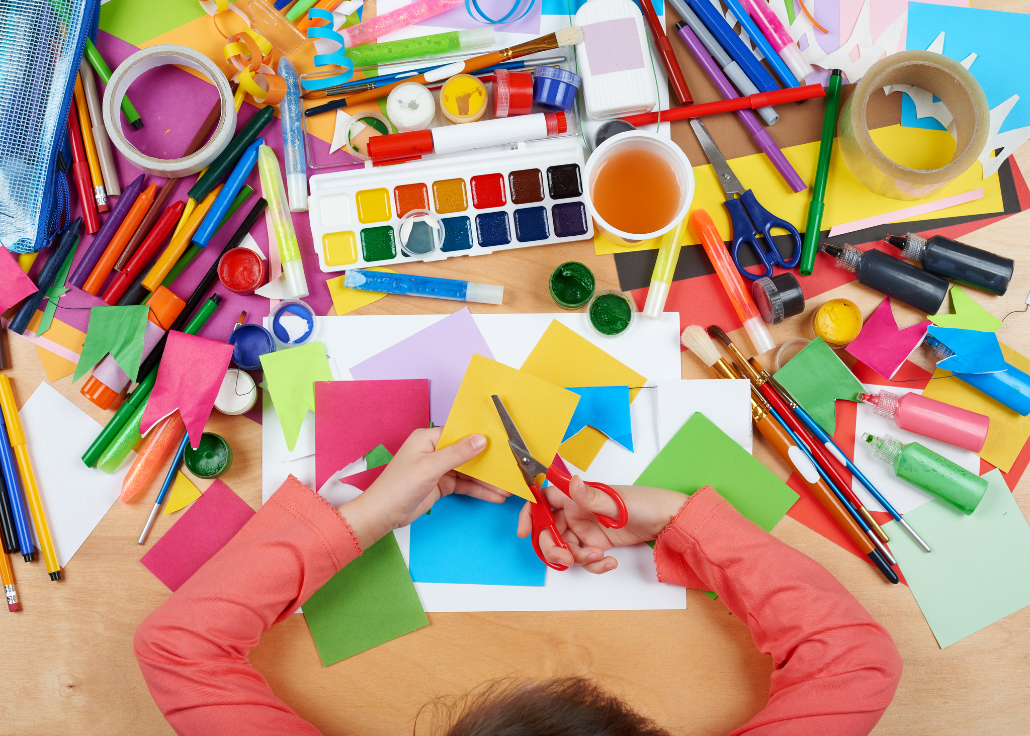 an overhead shot of a desk covered in colorful art supplies. A child's hands cut paper.
