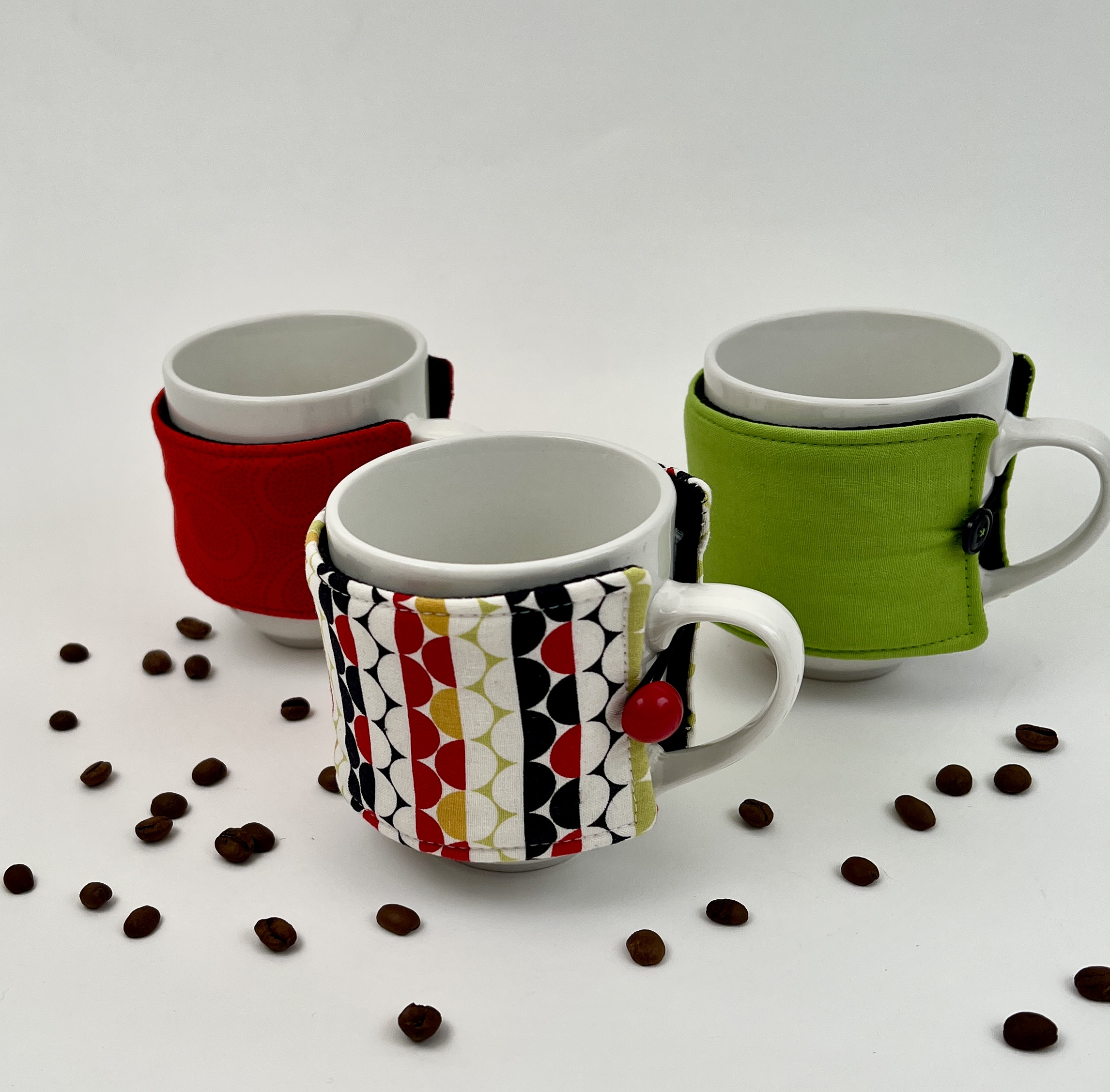 3 mugs with sewn fabric cozies and buttons on a white table with coffee beans scattered around them