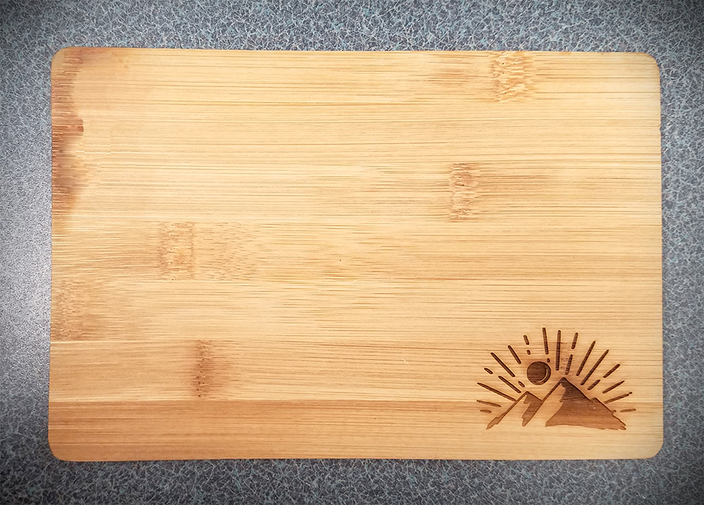 Bamboo cutting board with engraved mountain with sun behind it on bottom right corner