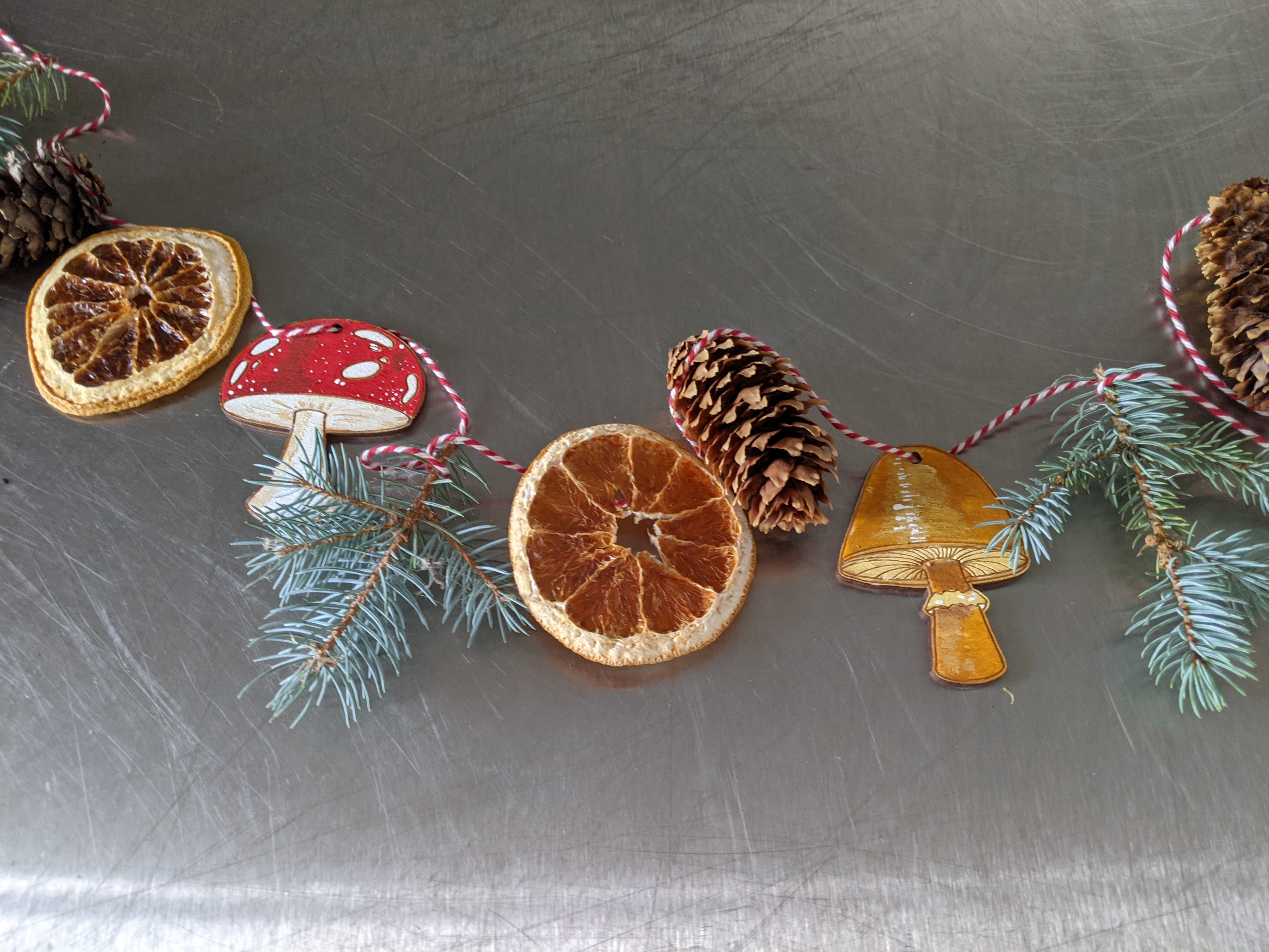 Garland made of dehydrated orange slices, pine cones, pine branches, and painted laser cut mushrooms on a metal table