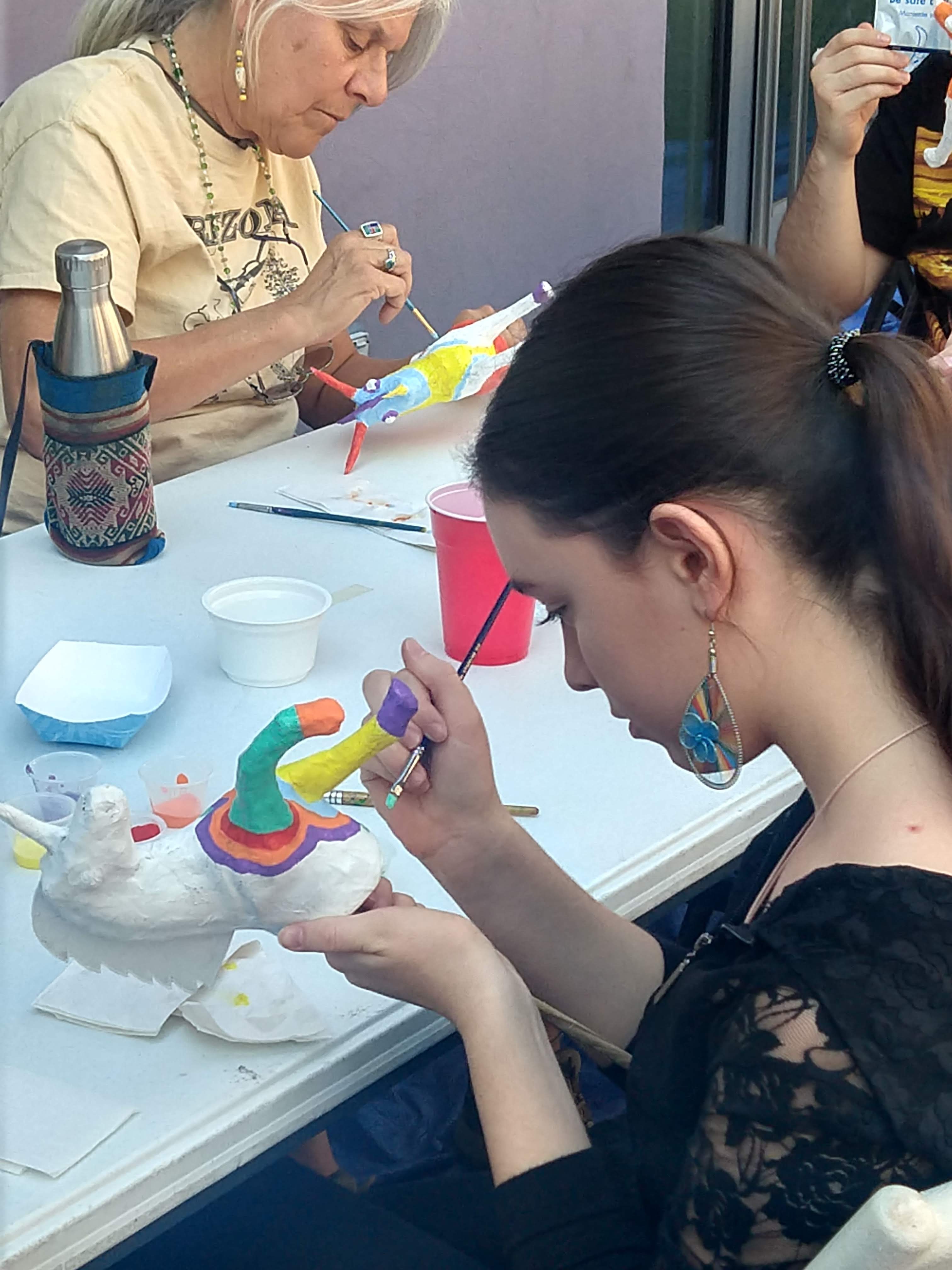 Two women, different ages, painting small alebrijes sculptures.