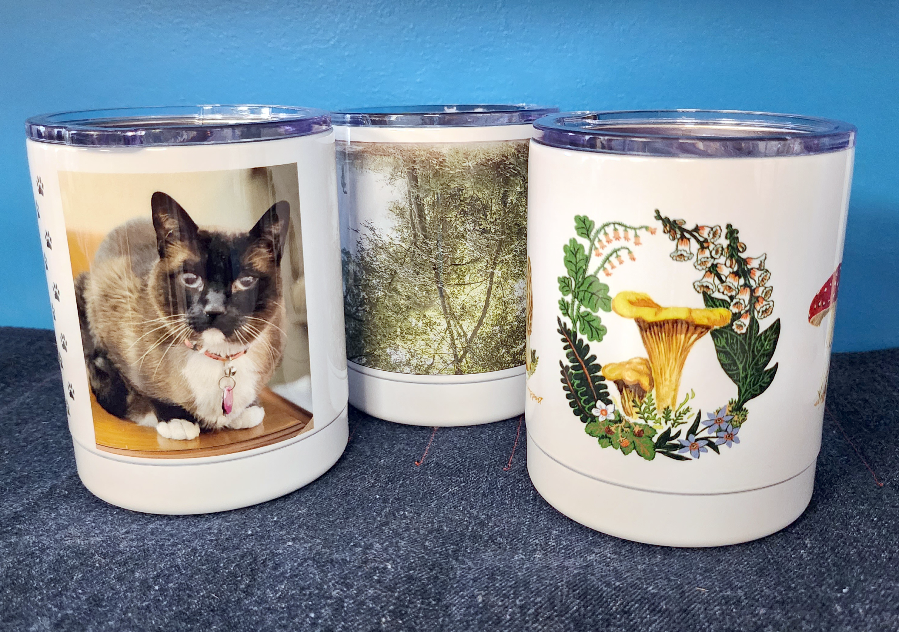 Three sublimation tumblers on a blue background printed with a cat, trees and mushroom illustration