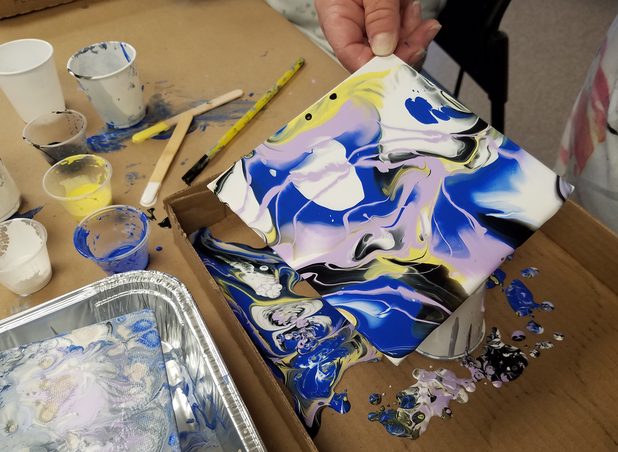 Hand holding a pour painting in progress with areas of blue, purple, yellow, and white paint mixing together. In the background are paint cups and supplies.