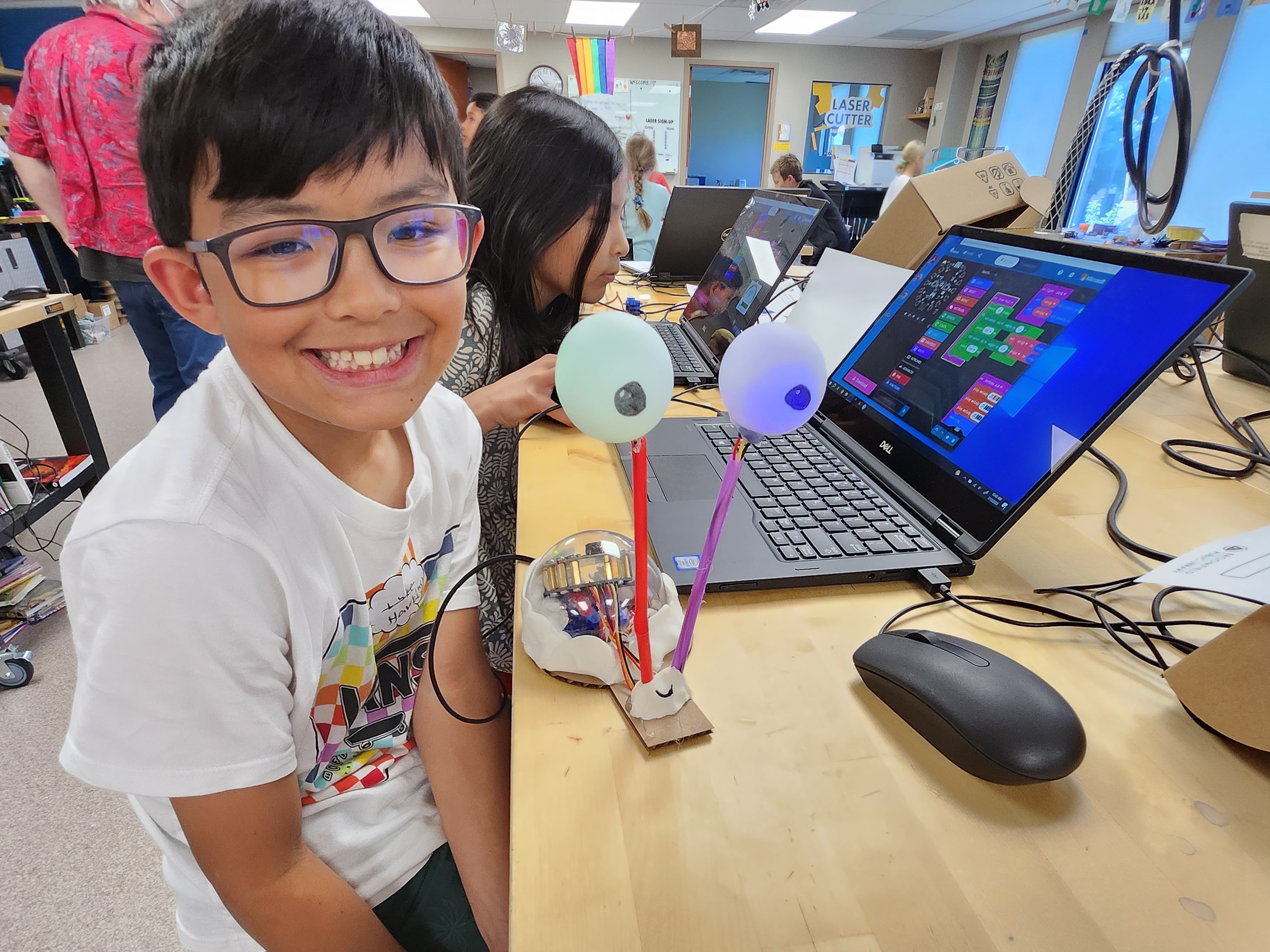 Tween poses at a table with his robot snail creation. A laptop behind the snail shows the code that makes it work.