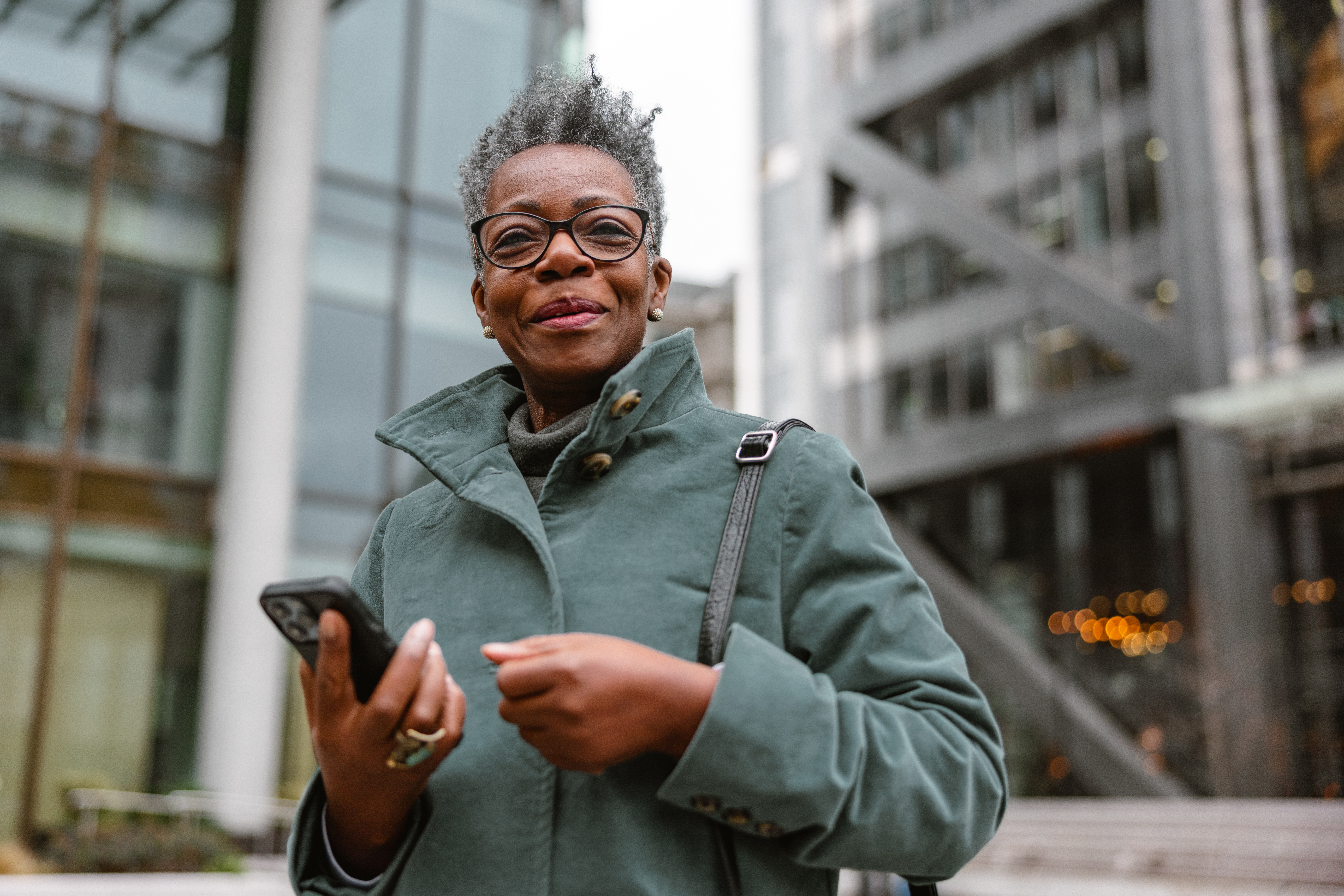 An older Black woman stands outside in a city holding a phone and smiling at the camera
