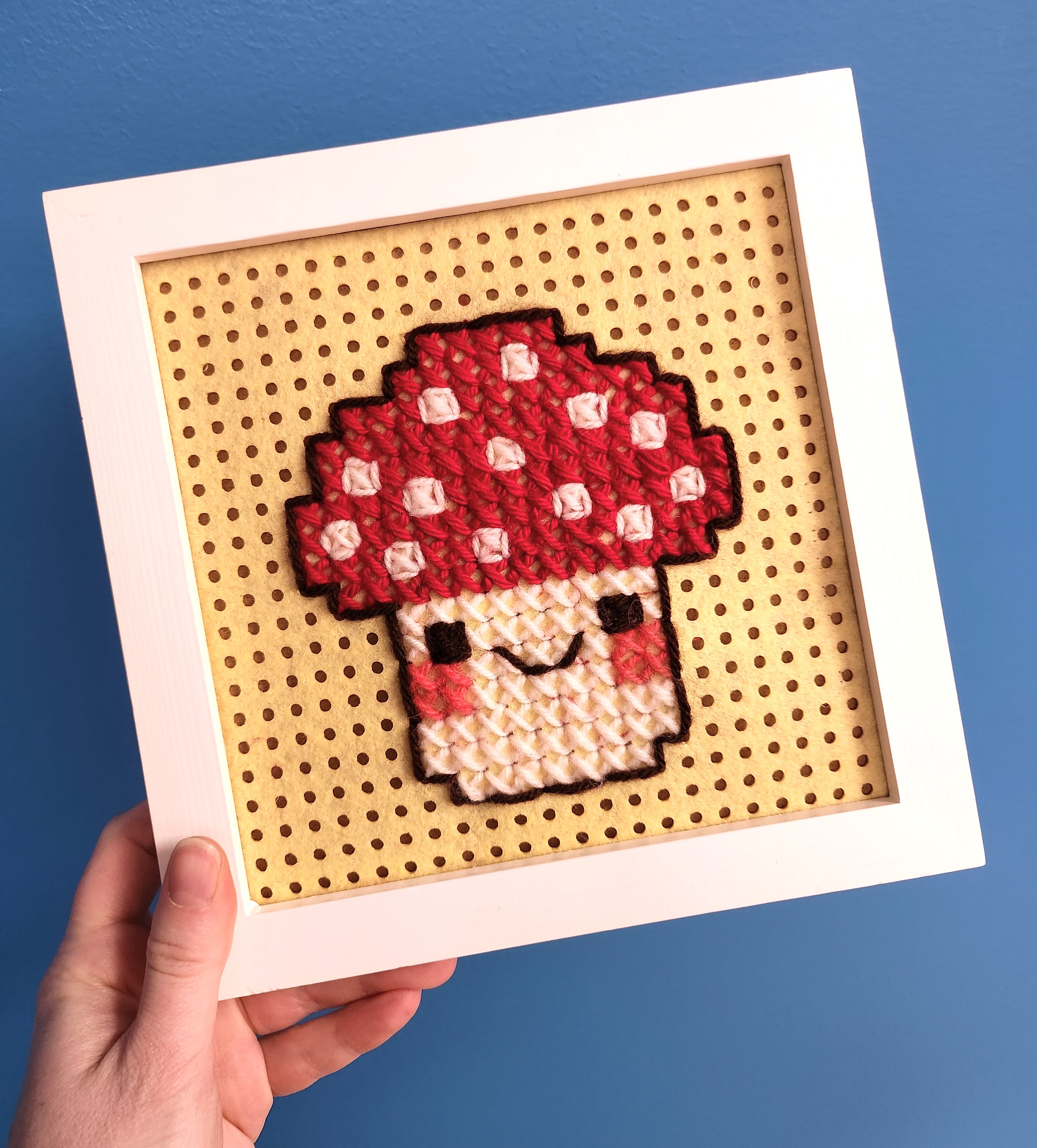 Hand holding a large cross stitch made with yarn in a wooden frame. The cross stitch is of a cute mushroom with eyes and a smiling mouth.