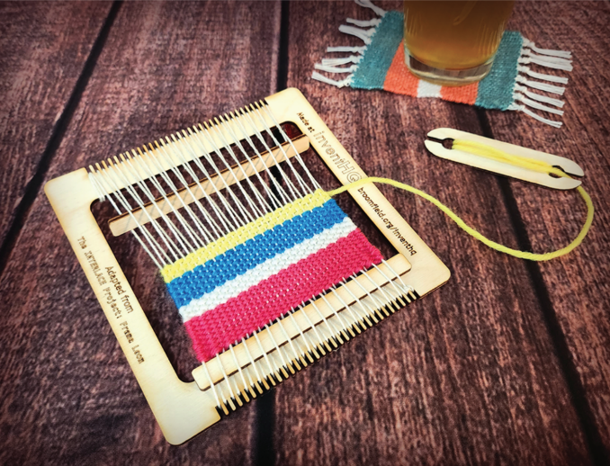 Laser cut loom with weaving project on a table with a beer glass in the background