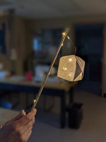 Hand holding wooden dowel wrapped in string lights with dangling white origami box glowing