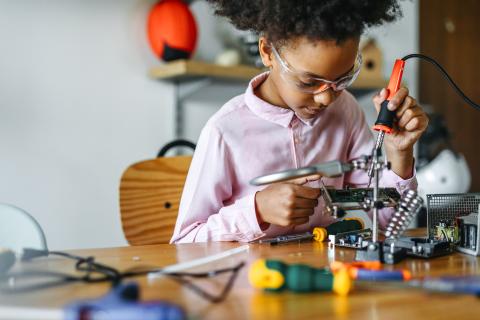 Girl at a table with safety glasses on working on a soldering project.
