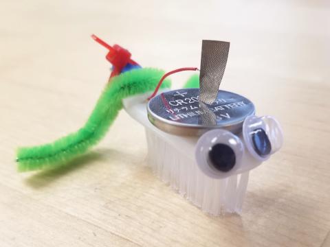 Bristlebot robot made from a toothbrush head, small motor, and battery with googly eyes and a green pipe cleaner tail
