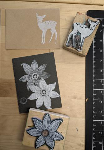 Block printing examples on table of a deer stamp on an envelope and flower stamp on a notebook