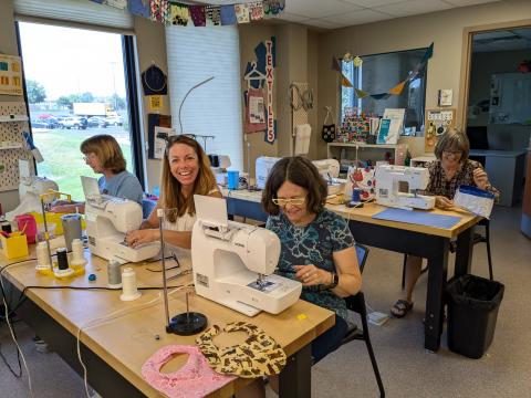 Three people sewing and one laughing in front of large windows. 