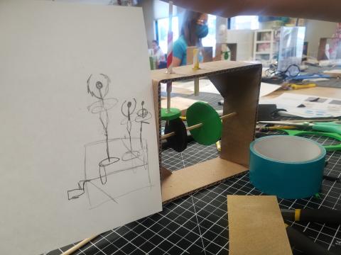Cardboard automata on a table with drawing representing it's theme next to it