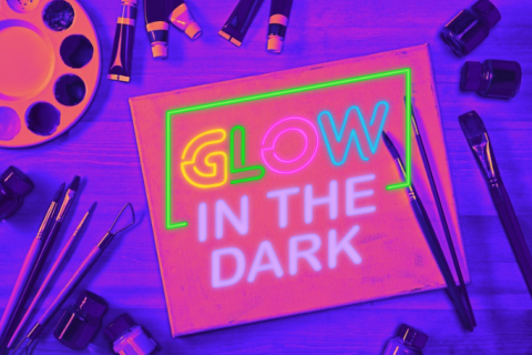 Glow in the dark sign superimposed over paper surrounded by paintbrushes, tinted to appear as if in blacklight