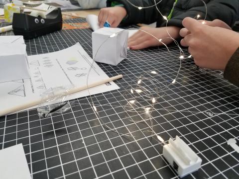 Hands working on paper crafts with string lights in the foreground