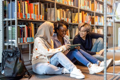 3 teens hanging out together, looking at an ipad on the floor of a library in front of a shelf of books