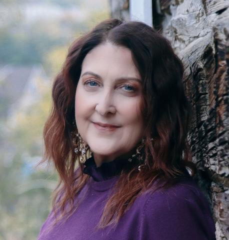 artist headshot woman facing the camera with red shoulder length hair wearing a purple turtlenec