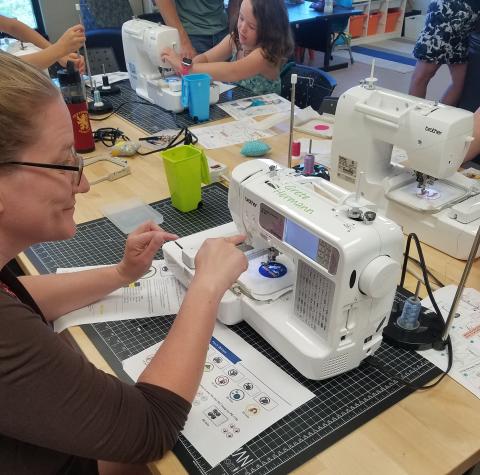 Person pressing a button on an embroidery machine. Behind them are more machines with people trying out embroidery.