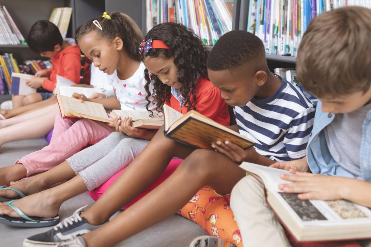 a line of children sit in a library reading books with their backs against a book shelf