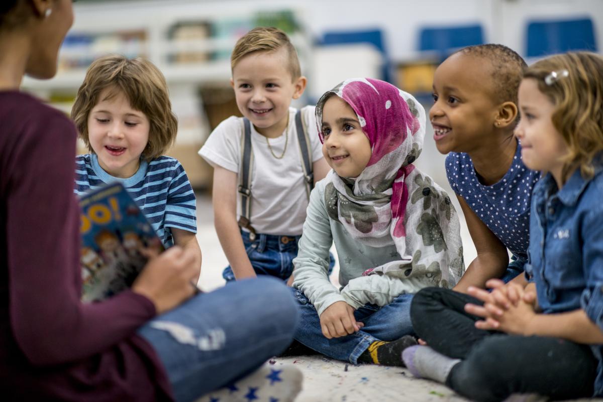 a group of five children listen eagerly to a story. A seated adult holding an open book is visible in the foreground