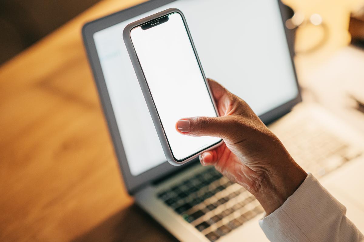 A hand holds a smartphone with a blank screen above a computer, also with a blank screen, on a table