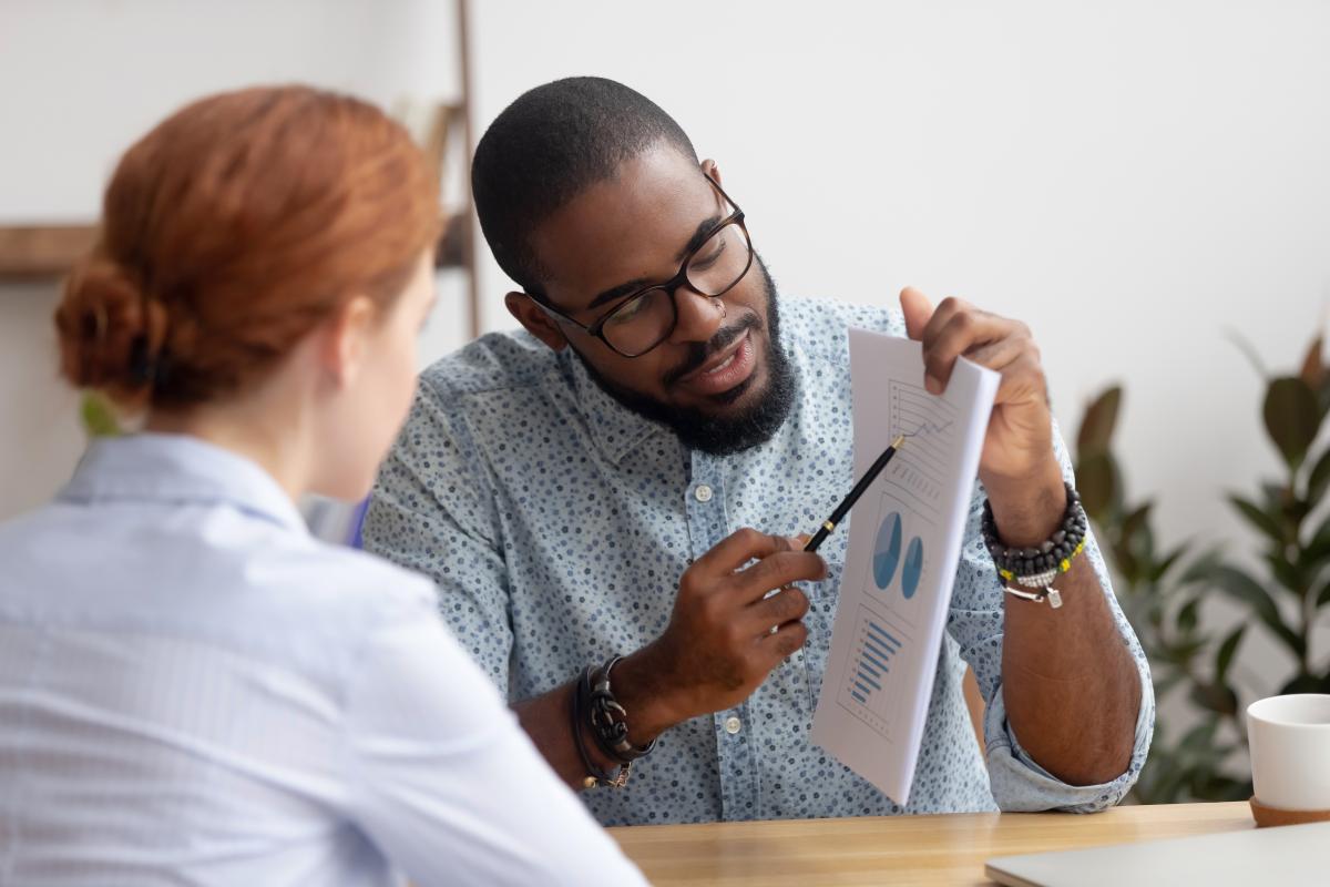 A man points out something on a sheet of paper with a pencil to a woman sitting across from him