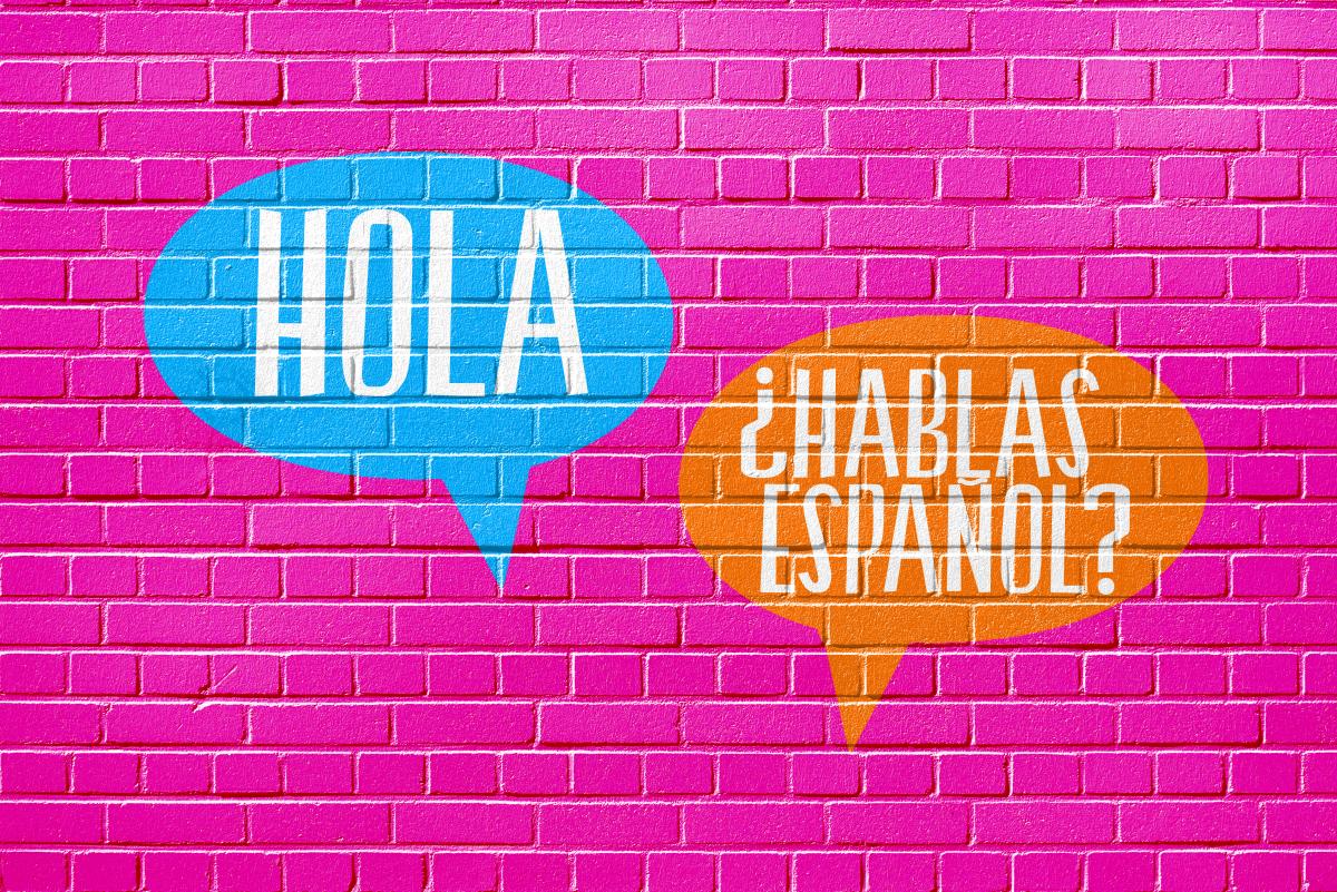 Two speech bubbles spraypainted onto a hot pink wall read "HOLA" and "¿HABLAS ESPAÑOL?"