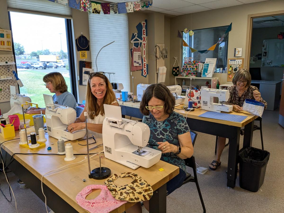 Three older adults using sewing machines and one laughing in front of large windows. 