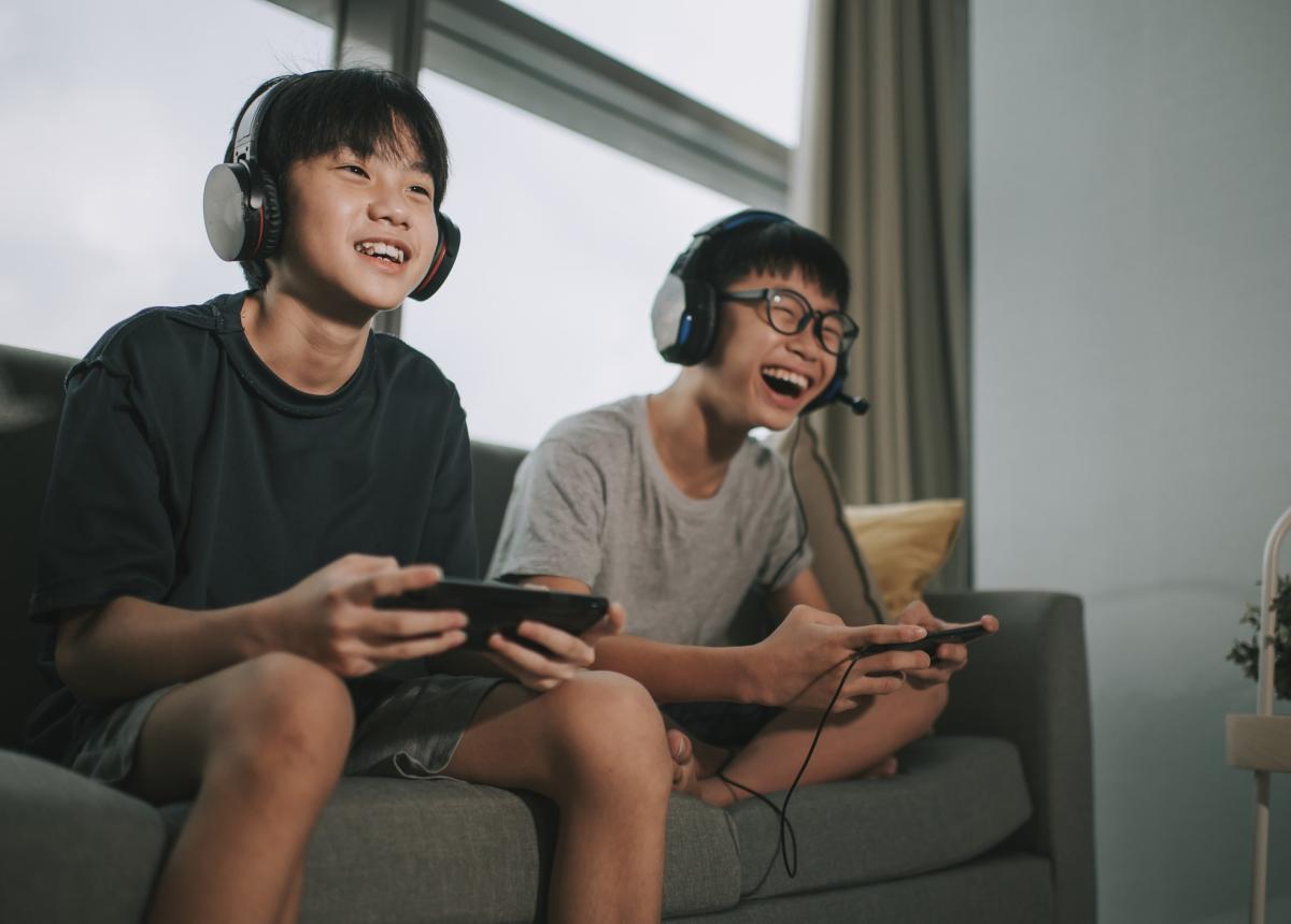 Two tweens sitting on couch, playing video games and laughing