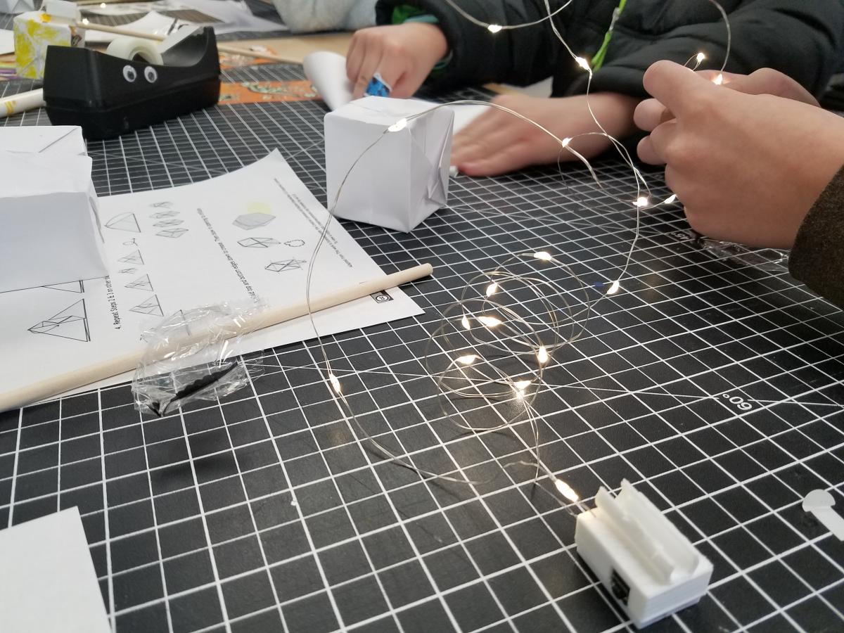 Hands working on paper crafts with string lights in the foreground