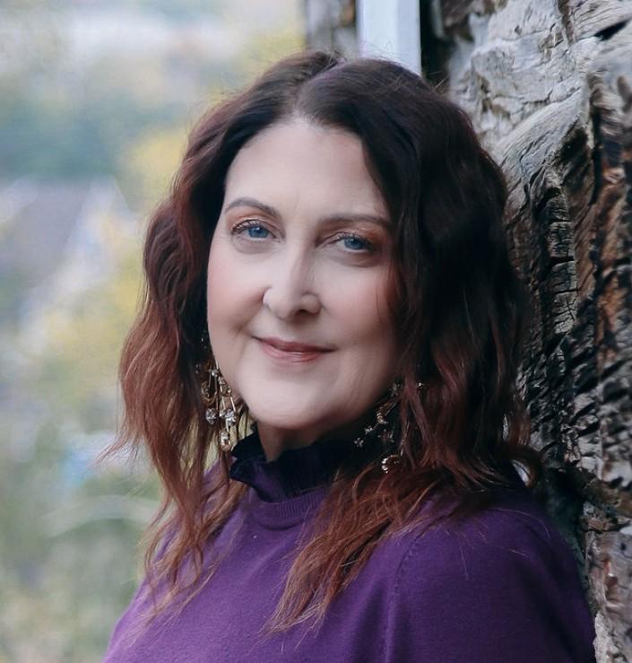 artist headshot woman facing the camera with red shoulder length hair wearing a purple turtlenec